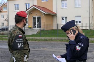 Umundurowana policjantka trzymająca telefon w ręku, obok żołnierz Żandarmerii Wojskowej.