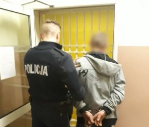 Policjant z osobą zatrzymaną, która ma zapięte kajdanki na rękach trzymanych z tyłu