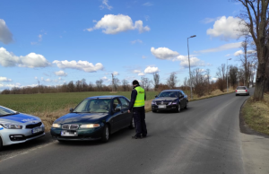 Na zdjęciu policjant badający kierującego pojazdem osobowym na zawartość alkoholu. Za nim stoi kolejny samochód. po lewej radiowóz policyjny.