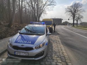 Na zdjęciu droga i policyjny radiowóz za którym policjantka legitymuje osobę w samochodzie.