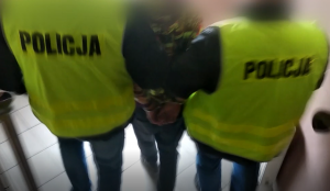 Na zdjęciu dwóch policjantów w kamizelkach prowadzi po schodach osobę, która ma zapięte ręce w kajdanki.