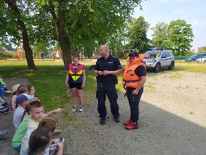 Na zdjęciu policjant tłumaczący uczniom zasady bezpiecznego wypoczynku nad wodą.