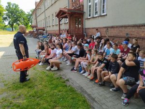 Na zdjęciu policjant tłumaczący uczniom zasady bezpiecznego wypoczynku nad wodą.